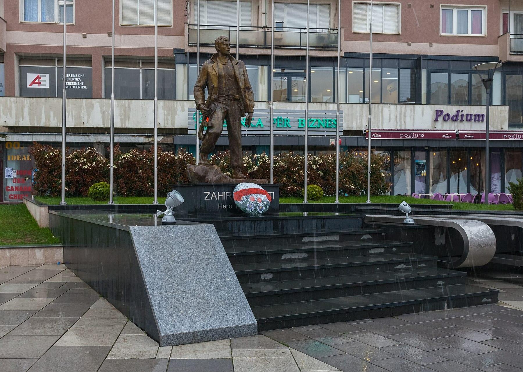 Bronze statue of a standing man on a black pedestal in front of apartment blocks.