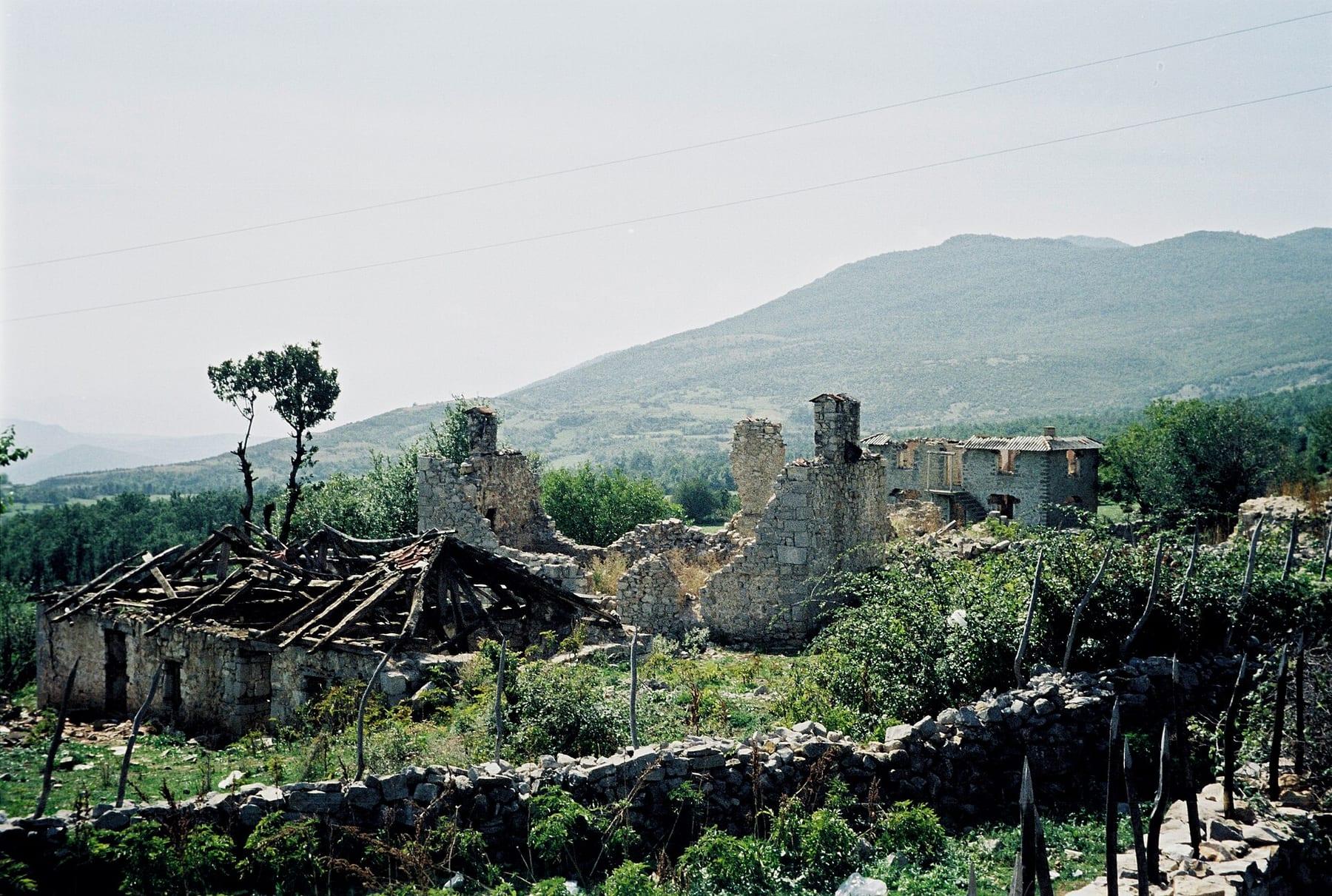Roofless ruined stone buildings among vegetation with a mountain behind.