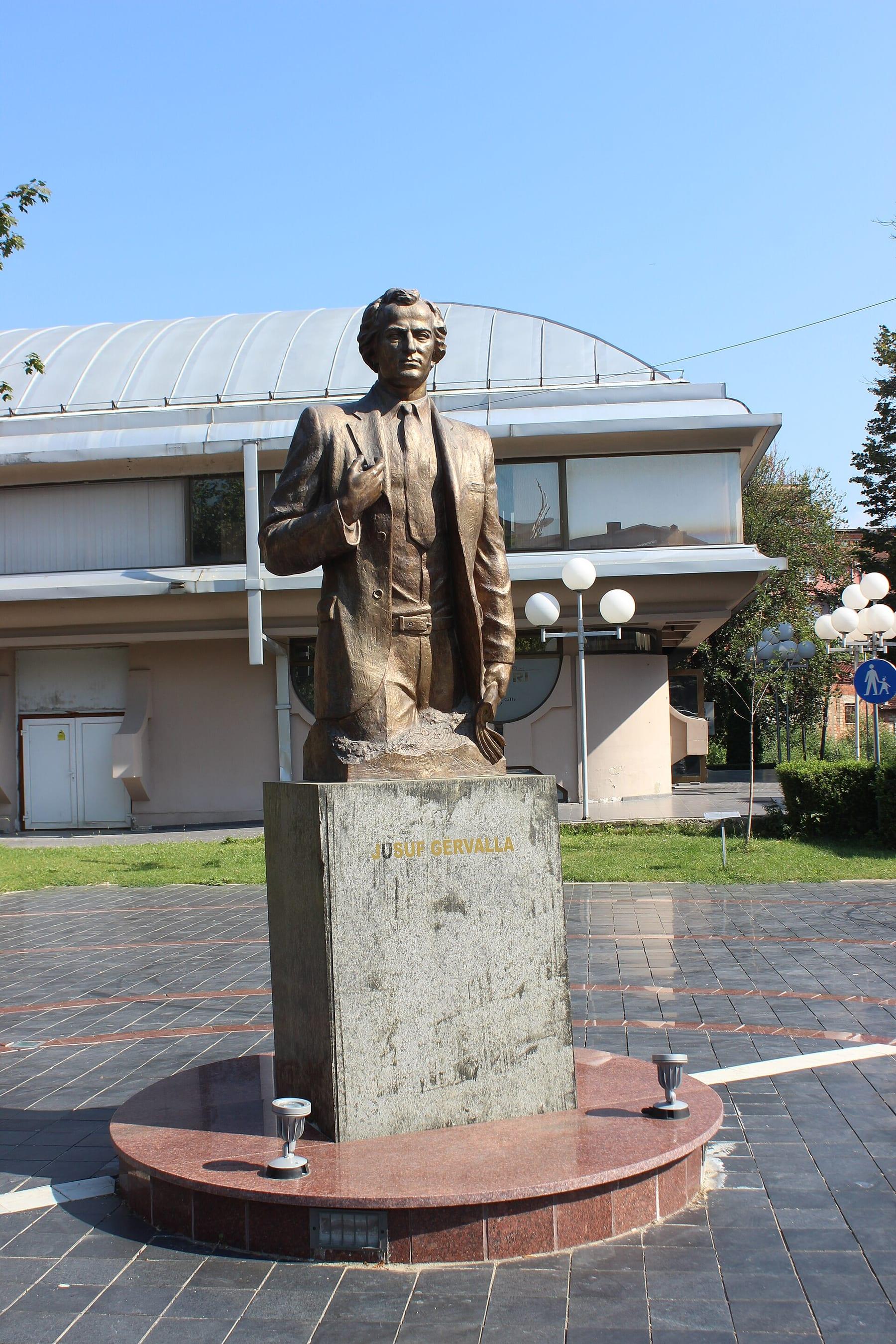 Gilded statue of a standing man on a stone plinth in front of a domed building.