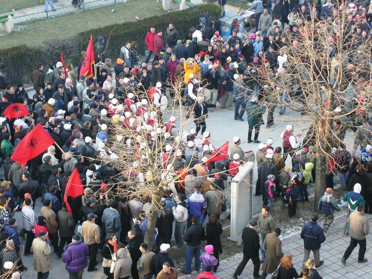 Elevated view of a dense crowd with red-and-black flags; several men in white plis caps.