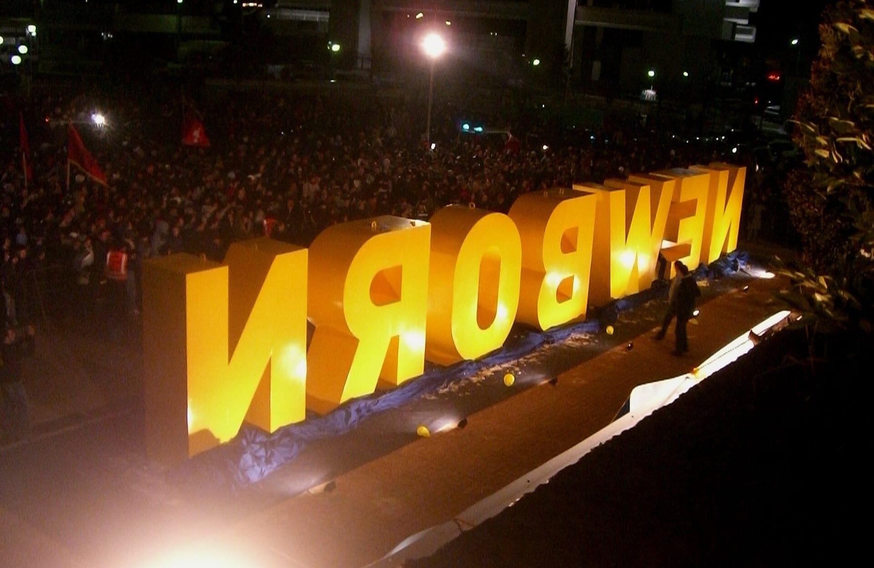 The yellow NEWBORN letters lit at night, surrounded by a large crowd.