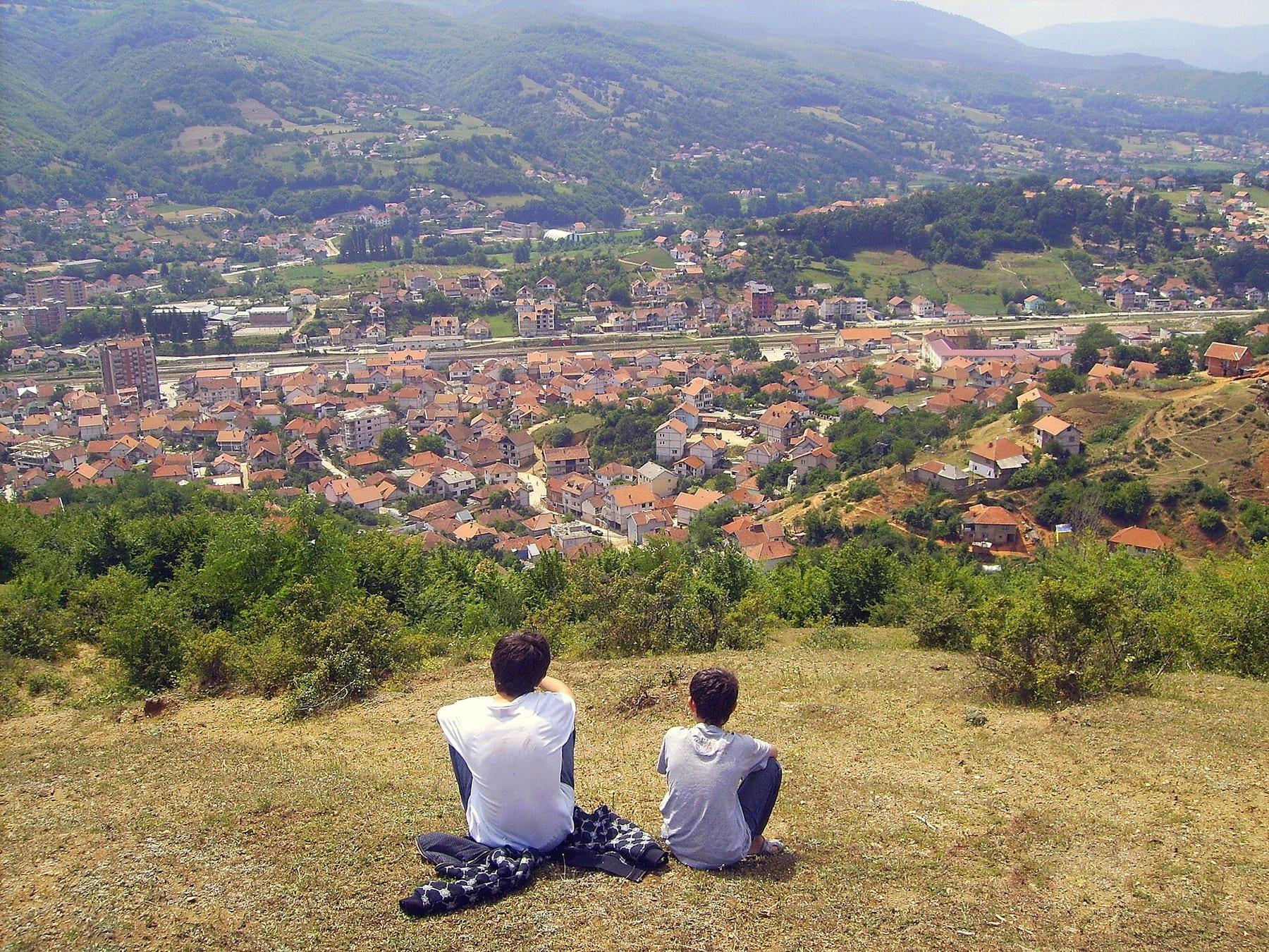 The town of Kaçanik with red roofs in a valley, seen from a hill; two people sitting with their backs to the camera.