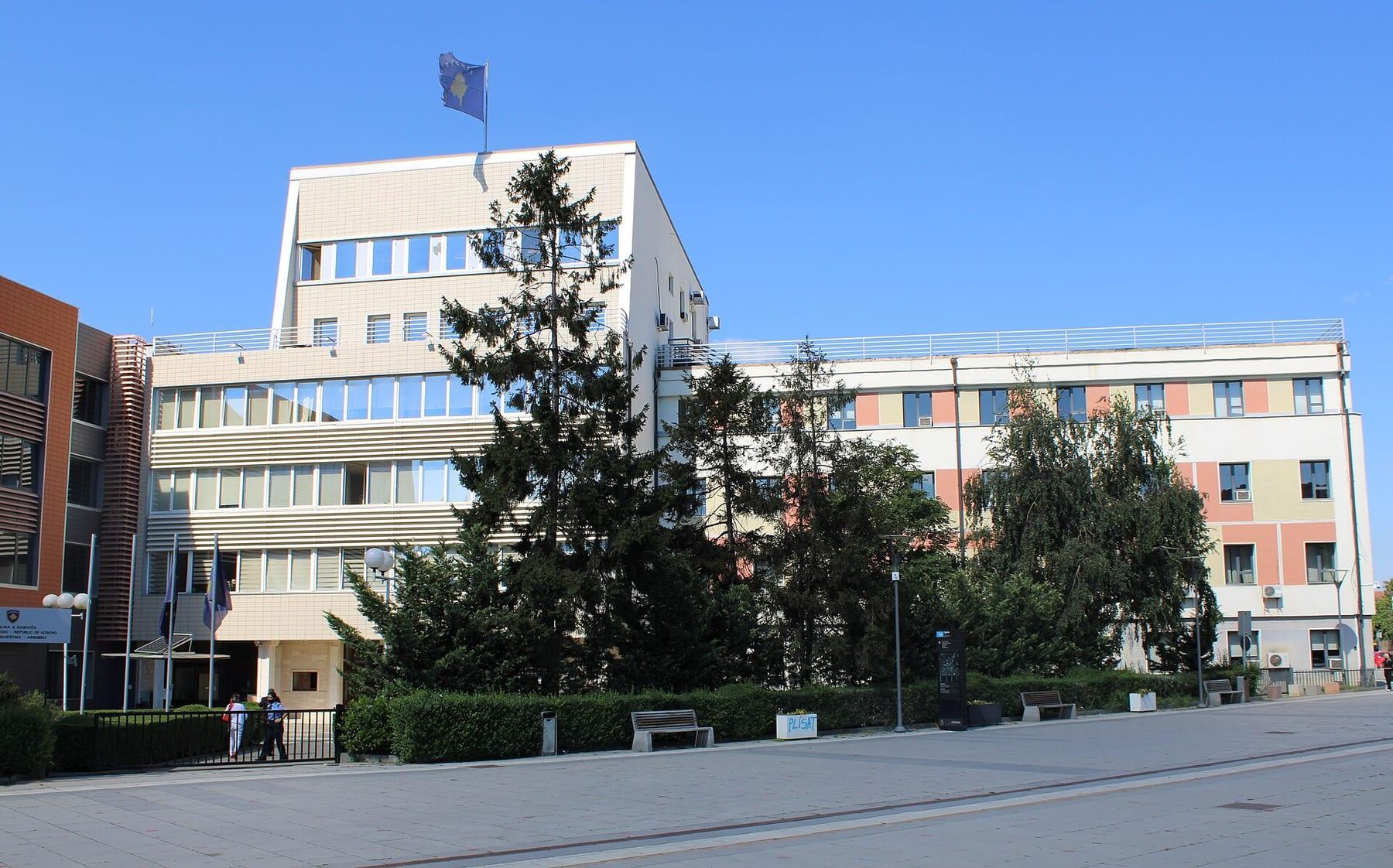 White and brick-coloured multi-storey building with a flag on top, behind a square with trees.