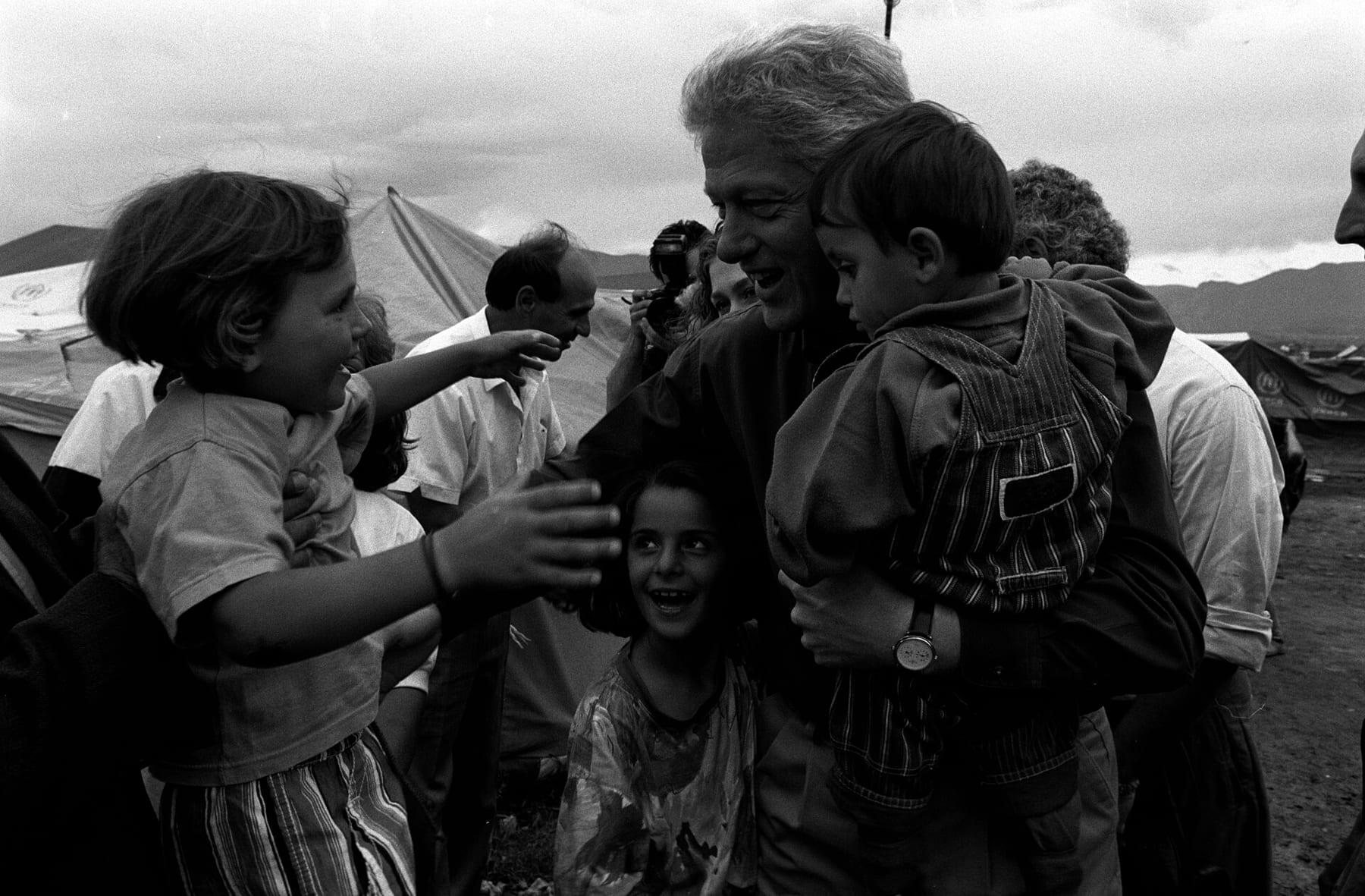 Black-and-white photograph: Clinton holds a child while other children reach towards him; tents behind.