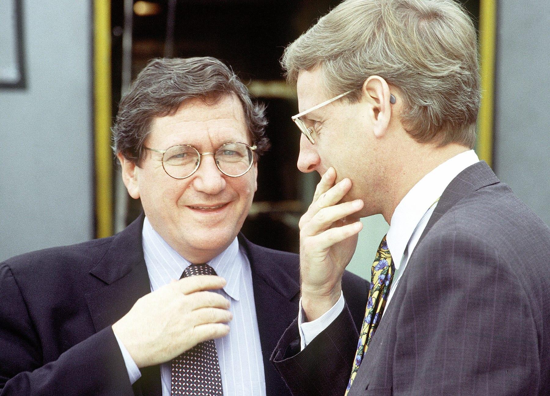 Two men in suits and glasses in close conversation; one is smiling.