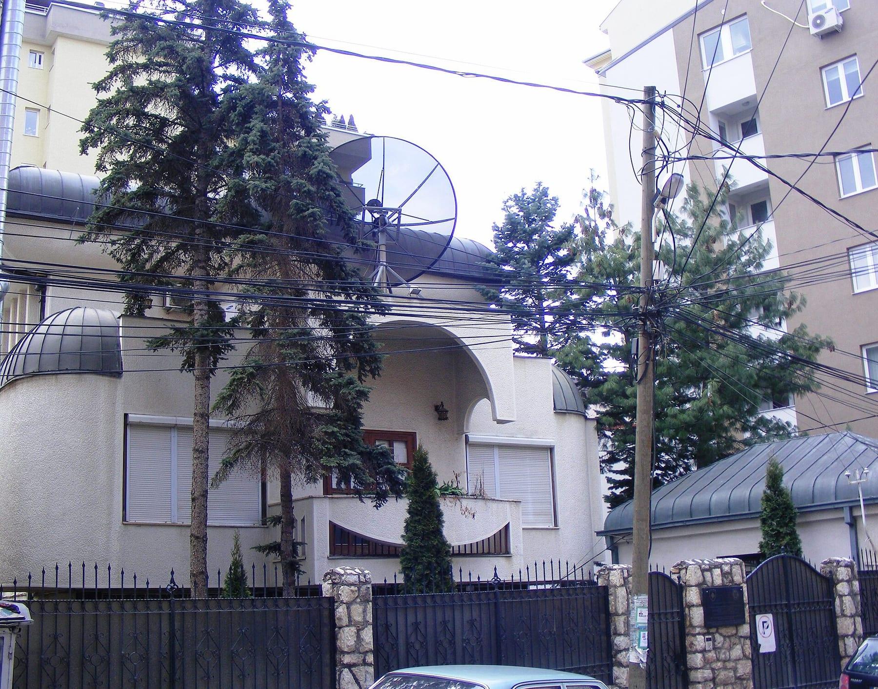 White two-storey house behind a metal fence, with fir trees in front.