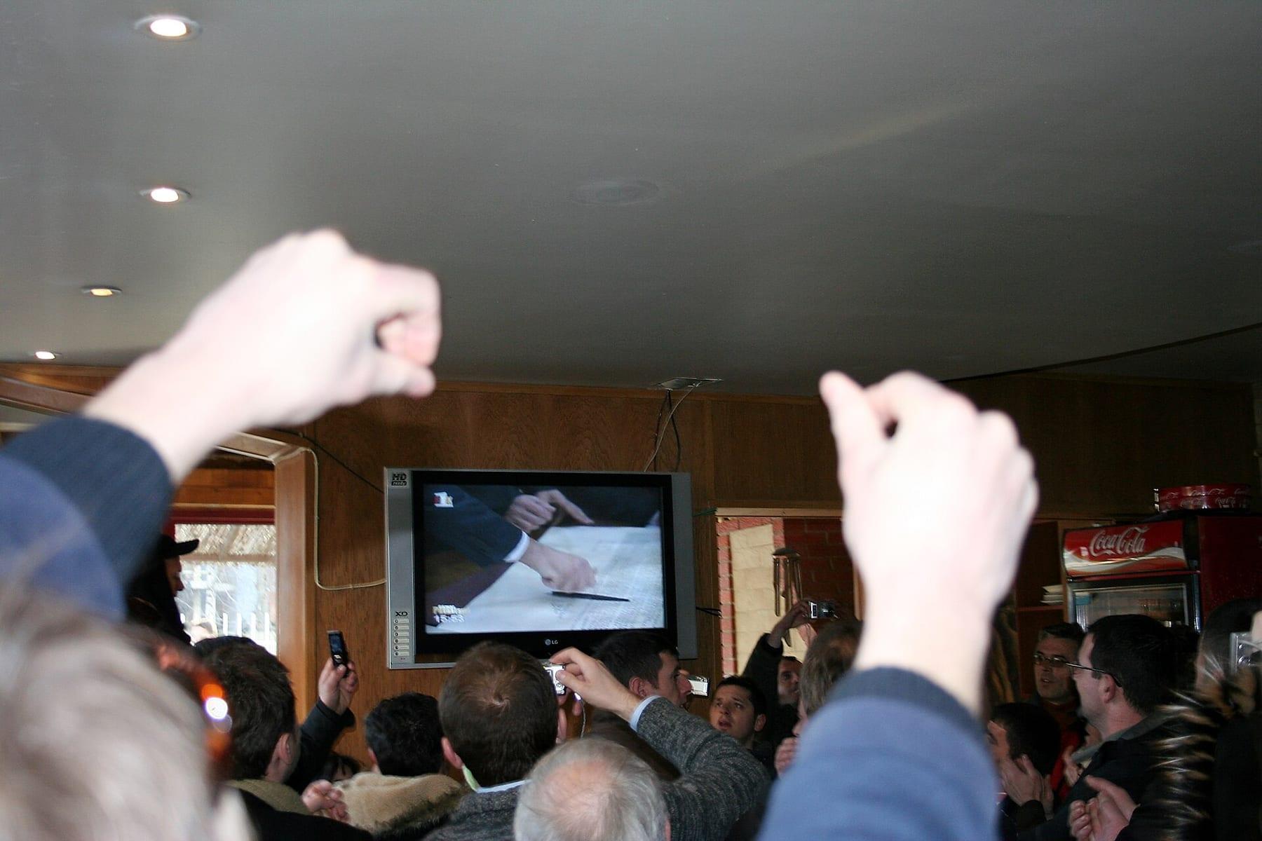 A crowd in a café with raised fists and phones in front of a television showing the signing.