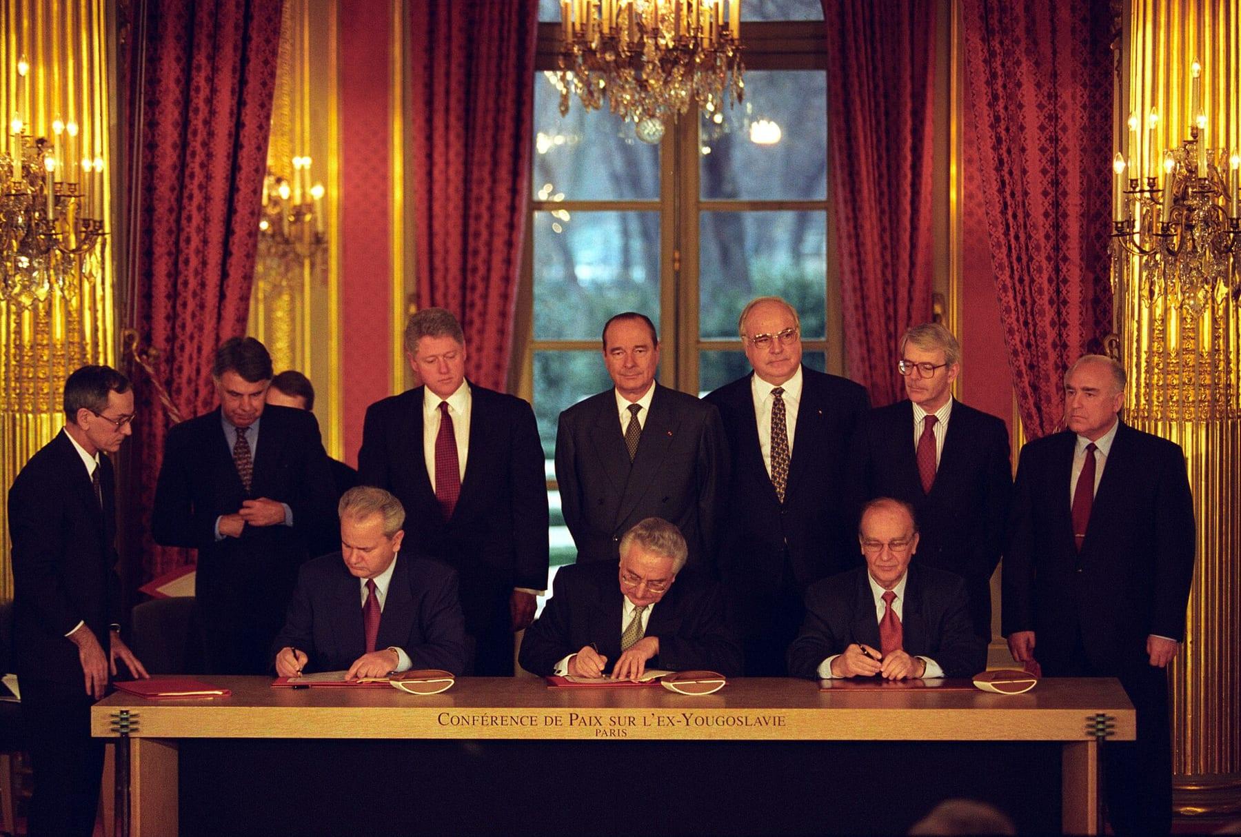 Three seated men signing at a long table, a row of heads of state standing behind them in a hall with red curtains.