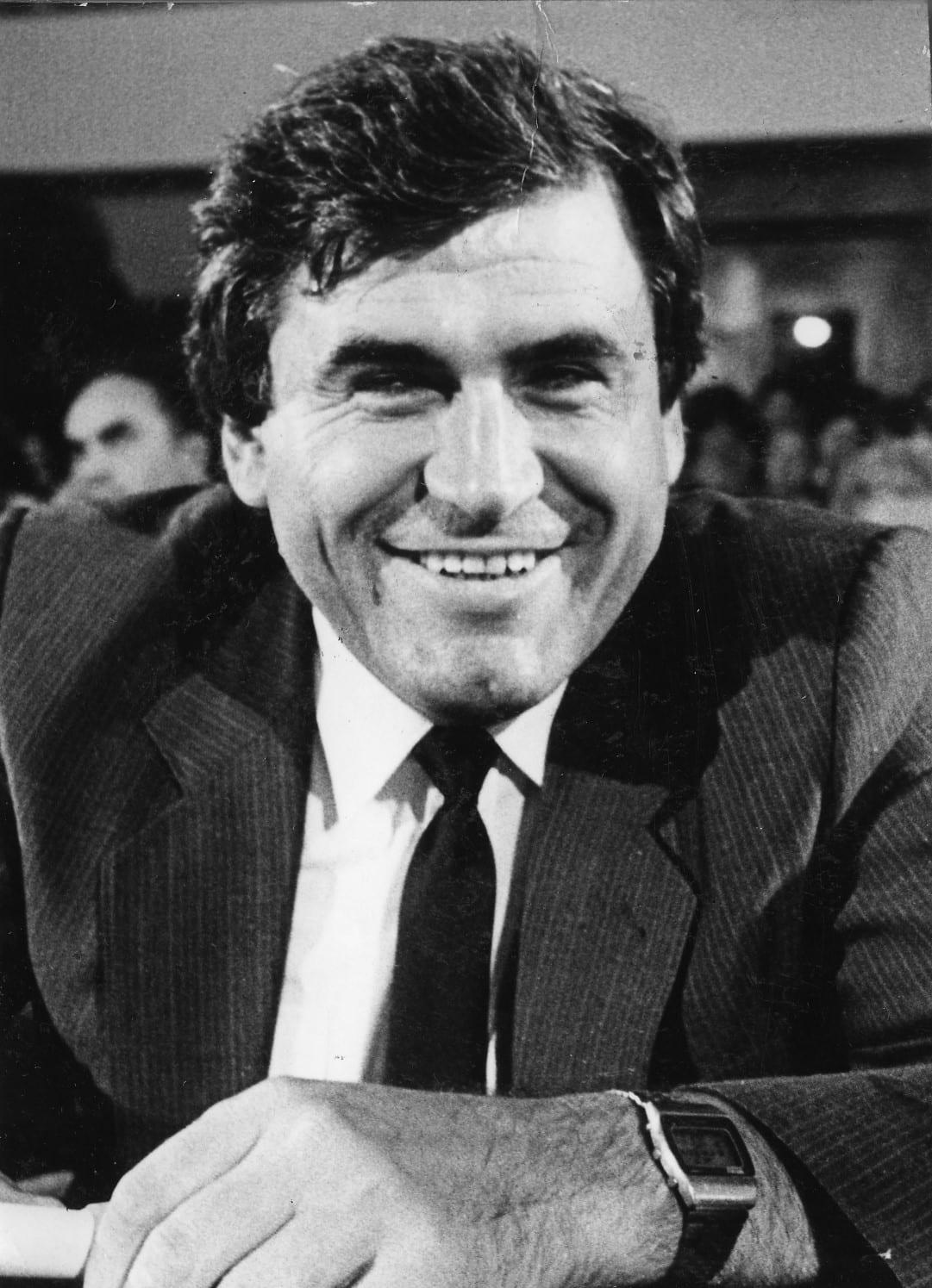 Black-and-white portrait of a smiling man in suit and tie, seated in a hall.