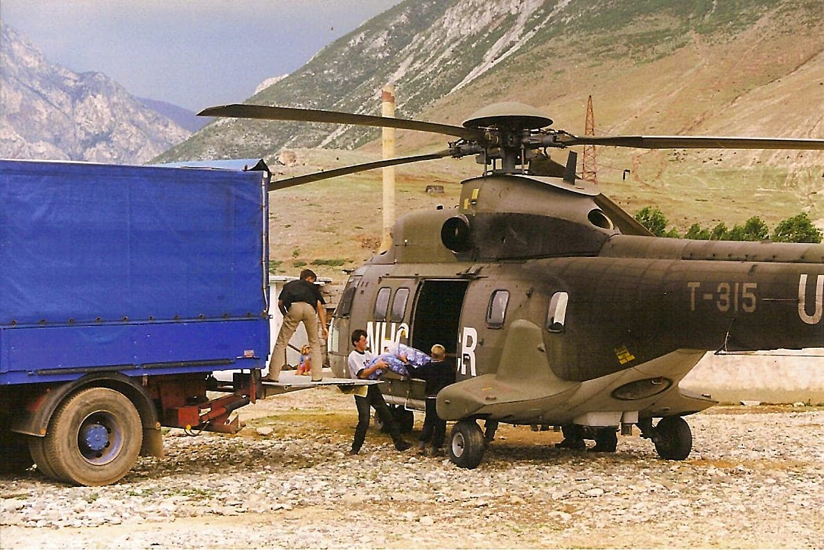 Military helicopter on the ground next to a blue-covered truck, mountains behind.