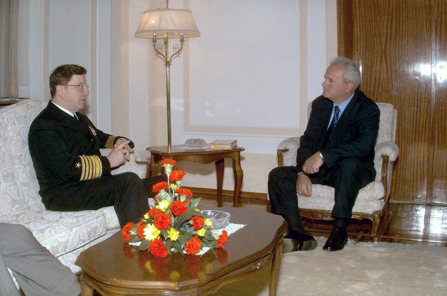 A naval officer in uniform and a man in a suit seated in armchairs in an official reception room.