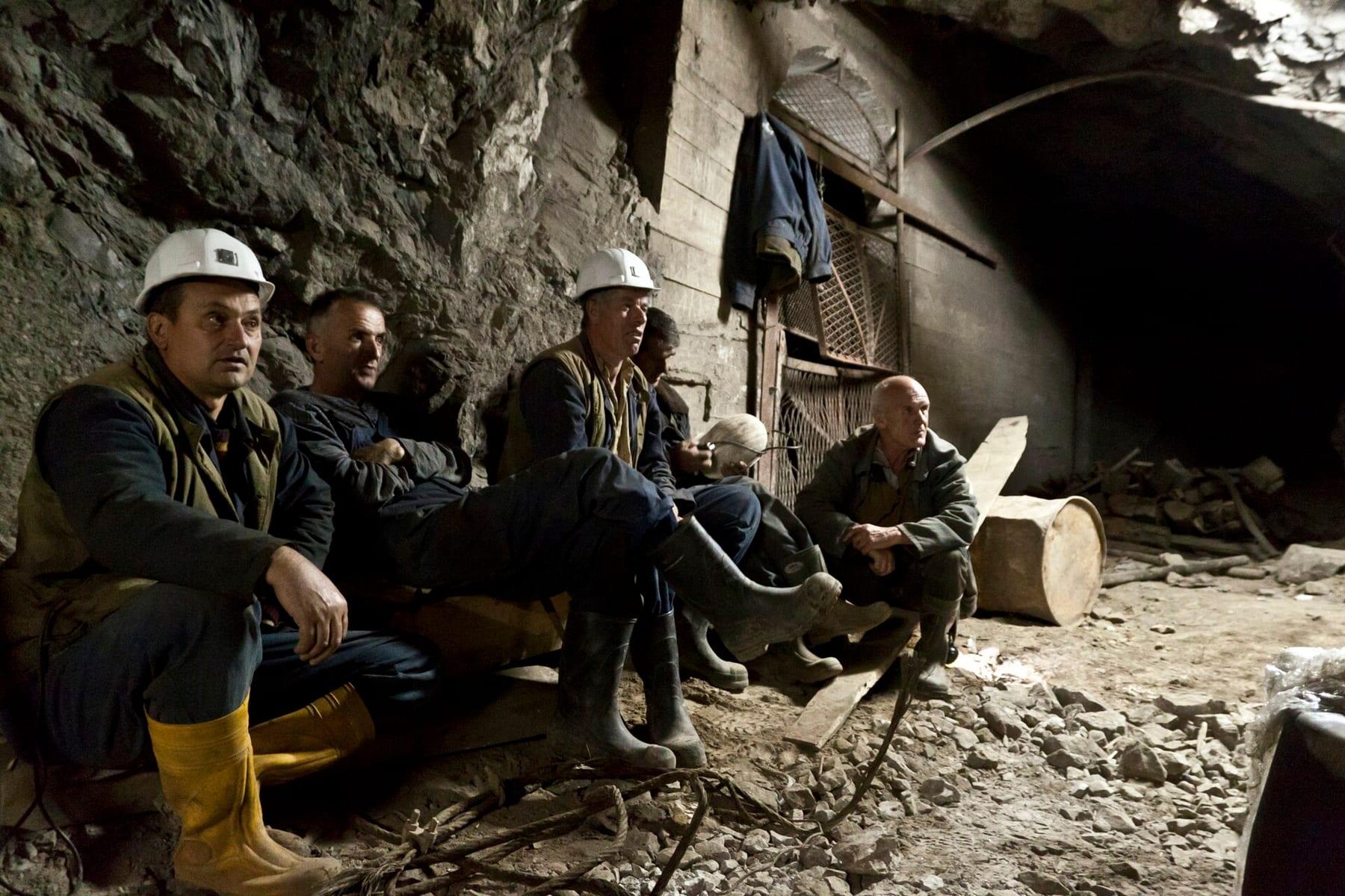 Four miners in white helmets and yellow boots sitting by the rock wall underground.
