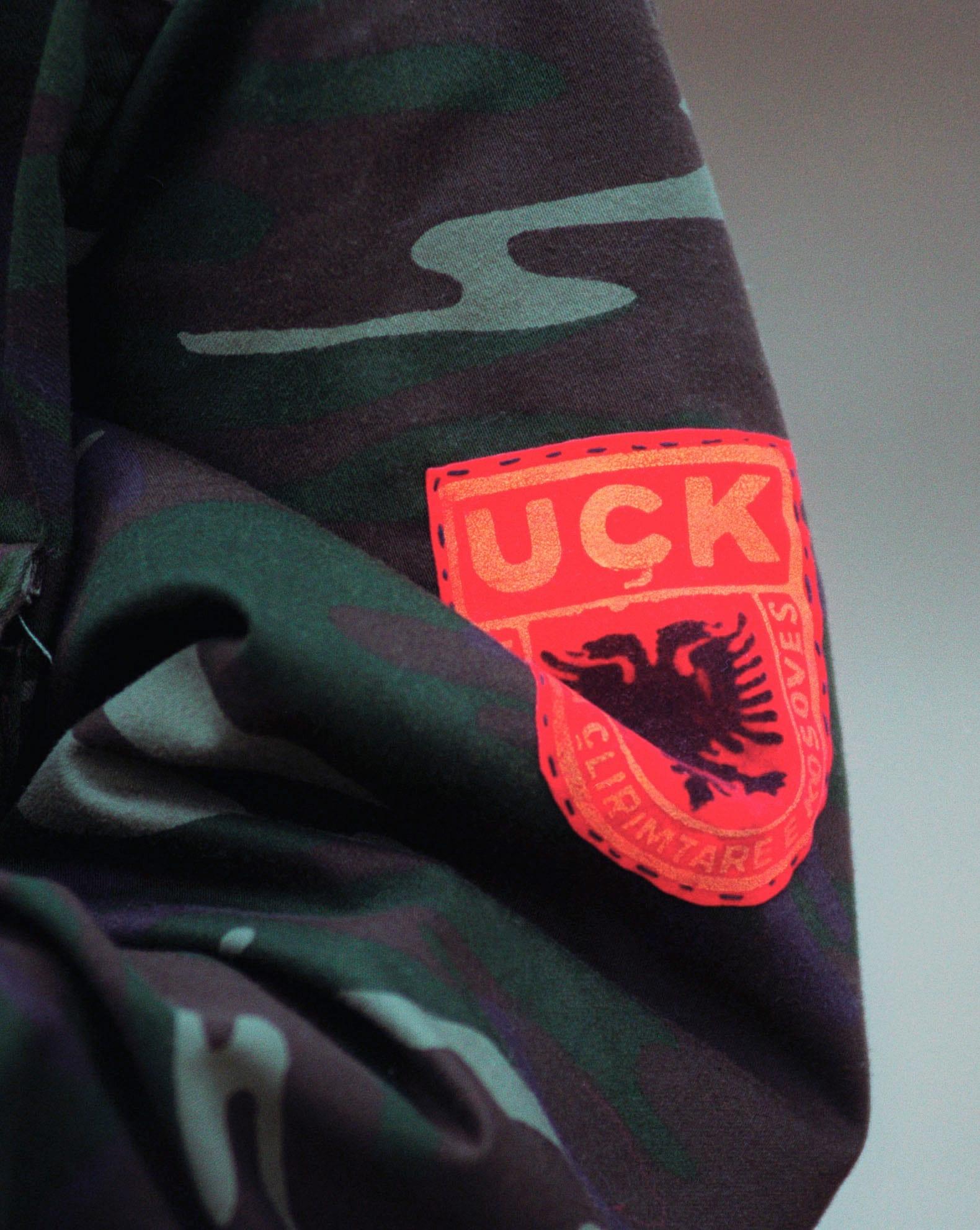 Close-up of a camouflage sleeve with a red patch bearing a black eagle and the letters UÇK.