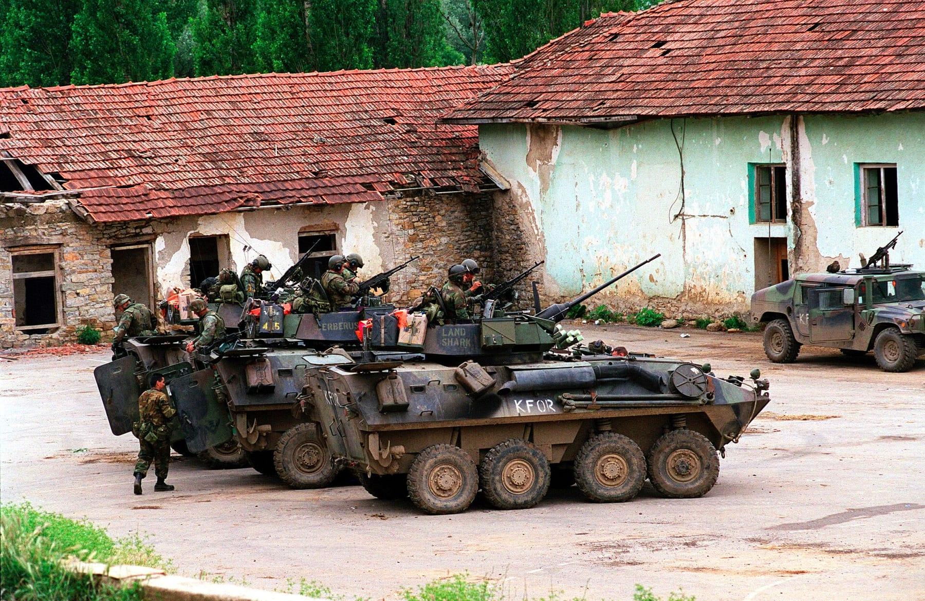 Wheeled armoured vehicles with Marines in front of damaged red-roofed houses in a village.