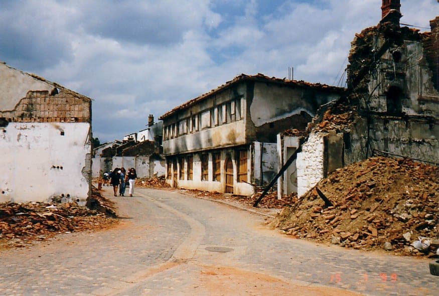 Cobbled street between burnt-out buildings and piles of rubble; a few pedestrians in the distance.