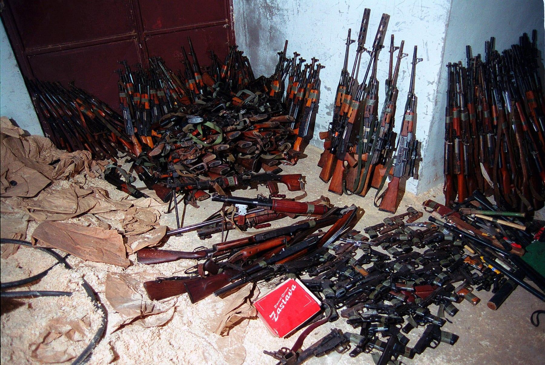 Pile of automatic rifles and other weapons leaning against a wall and lying on the floor.
