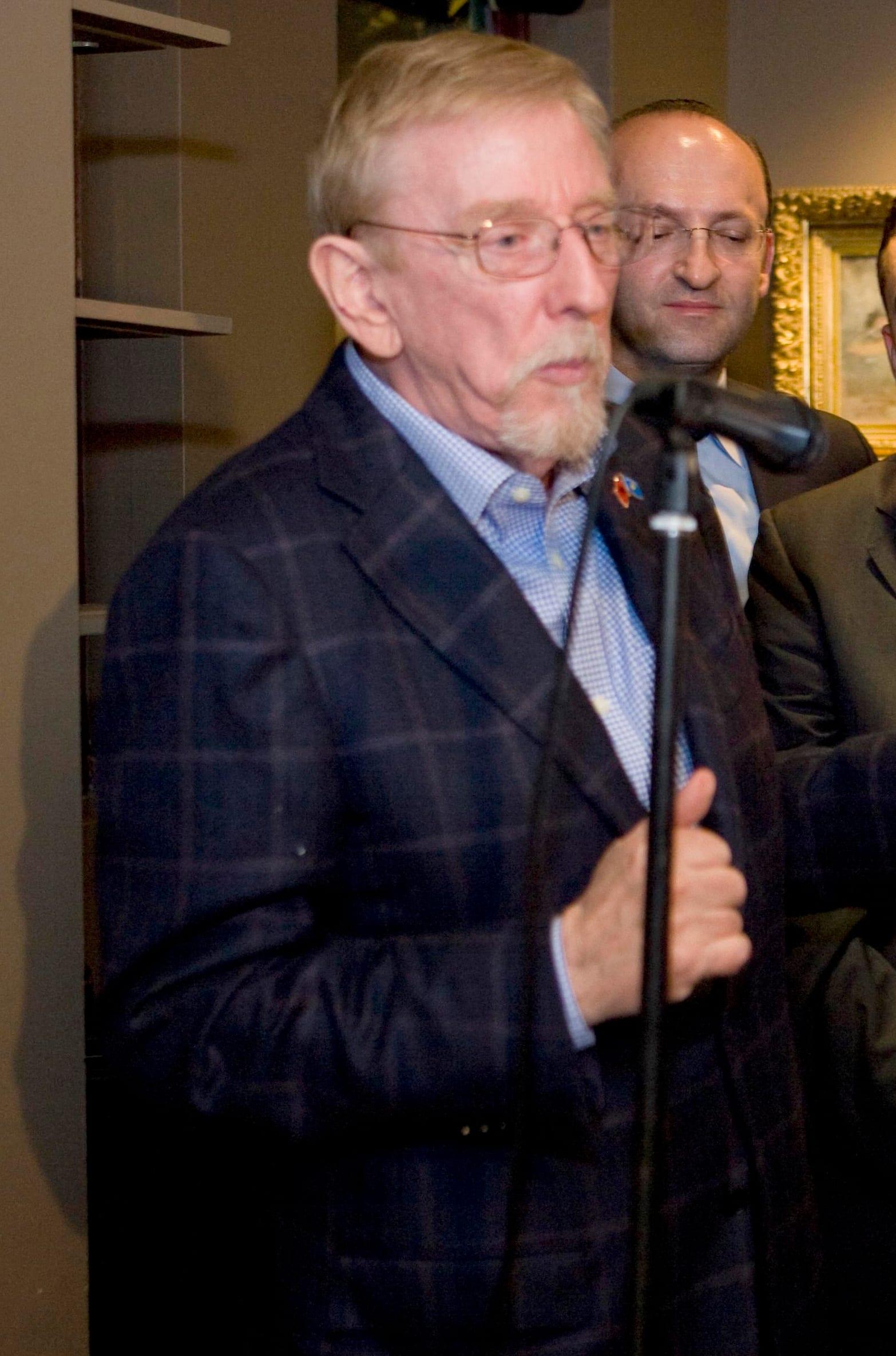 Older man with glasses and a short beard speaking into a microphone; another man behind him.