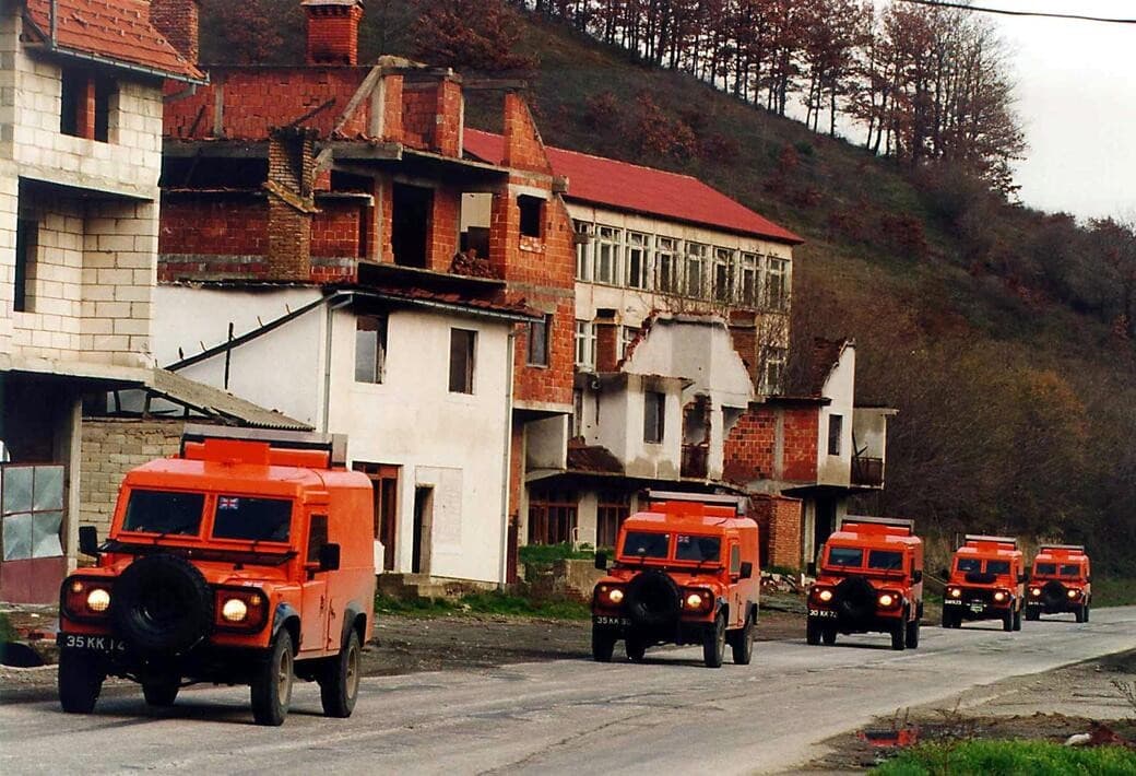 A column of orange armoured vehicles on a road beside damaged houses