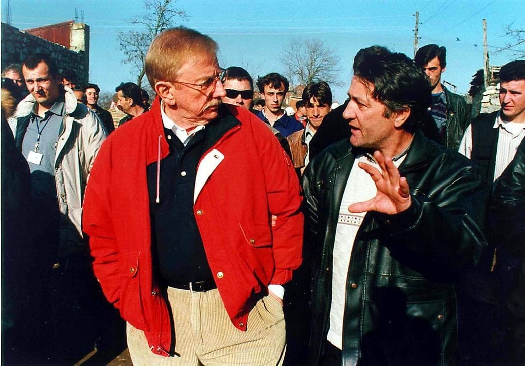 A man in a red jacket listens to a resident speaking, surrounded by people