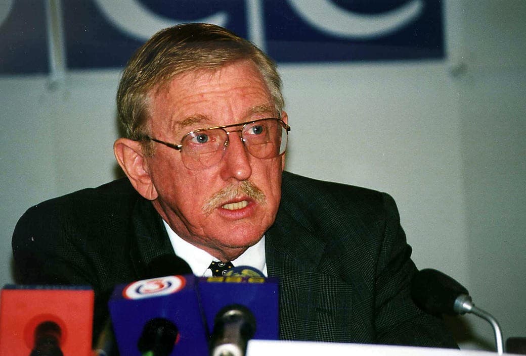 A man with glasses and a moustache speaks into microphones, the OSCE logo behind him
