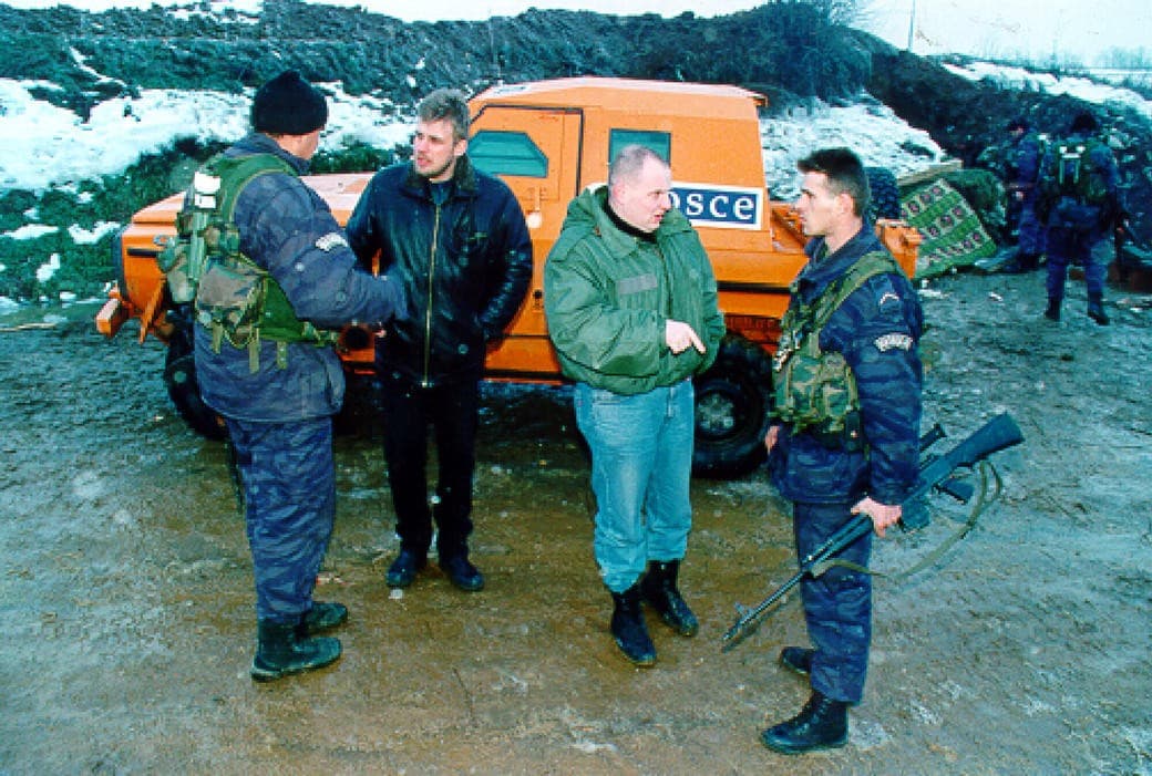 Civilian verifiers talk with armed policemen in front of an orange OSCE vehicle in the snow