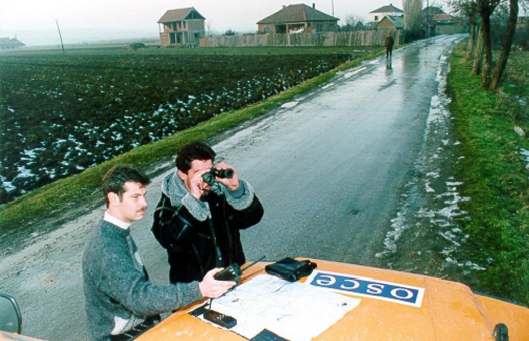 Two men beside an orange OSCE vehicle; one looks through binoculars along a village road
